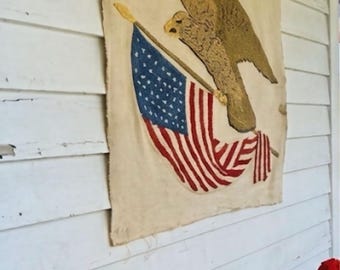 May include: A rectangular textile wall hanging featuring a textured, hooked-rug style illustration of a brown eagle in flight clutching a United States flag with red, white, and blue colors against a light beige, off-white fabric background.