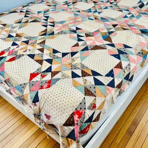 May include: A handmade quilt with a starburst pattern in a variety of colors, including pink, blue, red, and brown. The quilt is on a white bed frame and has a cream-colored background with small dotted patterns.