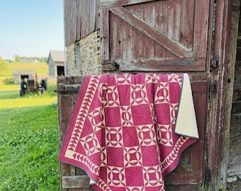 May include: A handmade quilt with a geometric pattern in shades of burgundy and cream, draped over a rustic wooden structure. The quilt features a repeating design of interlocking circles and squares, with a triangle border. The background includes a barn and green grass.