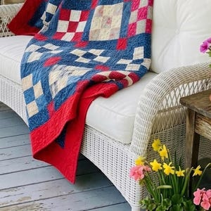Puede incluir: Un edredón hecho a mano con un diseño de patchwork en rojo, blanco y azul, sobre un sofá de mimbre blanco. El edredón presenta un patrón de cuadrados repetidos. La escena está ambientada al aire libre con flores en macetas.