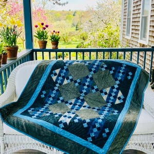 May include: A blue and green quilt with a geometric pattern rests on a white wicker couch on a porch. The quilt features squares of varying shades of blue and green, with a blue border. The porch overlooks a garden.