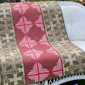 May include: A rectangular quilt featuring a central vertical panel of pink and dark red geometric star patterns flanked by two side panels of brown and beige floral and leaf prints, all finished with a solid brown fabric border trim.
