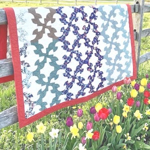 May include: A rectangular quilt featuring a repeating curved geometric pattern in shades of brown, green, blue, and white, finished with a solid red fabric border. The quilt is draped over a wooden fence above a garden of colorful tulips and daffodils.