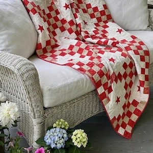 May include: A quilted throw blanket featuring a red and beige checkered pattern with scattered red star appliques. The fabric has a visible stitched texture throughout, creating a grid-like design that covers the surface of the textile piece.