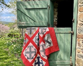 Puede incluir: Un edredón hecho a mano con un borde rojo y patrones geométricos en tonos de gris, rosa y azul marino está colgado sobre una puerta verde desgastada. El edredón se exhibe al aire libre, con un granero rústico y exuberante vegetación en el fondo.