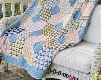 Könnte beinhalten: Eine bunte Patchwork-Decke mit geometrischen Mustern in Blau, Rosa, Gelb und Grau, drapiert über einem weißen Korbstuhl. Die Decke liegt auf einer Holzveranda mit Blumen im Vordergrund, was eine gemütliche Szene schafft.