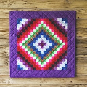 May include: A square quilt featuring a vibrant, multi-coloured geometric pattern of small squares arranged in concentric diamond shapes. The centre is a deep purple square, surrounded by layers of blue, green, red, and white, all framed by a purple border.