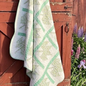 May include: A quilted blanket featuring a repeating pattern of white fabric blocks with green leaf-shaped motifs. The quilt is bordered and sectioned by solid light green fabric strips, showcasing detailed stitching throughout the entire textile surface.