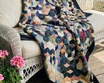 Könnte beinhalten: Eine handgefertigte Steppdecke mit einem geometrischen Muster in Blau-, Grün-, Rosa- und Cremetönen. Die Steppdecke ist über ein weißes Korbmöbel auf einer Veranda drapiert. Die Steppdecke hat einen dunkelblauen Rand.
