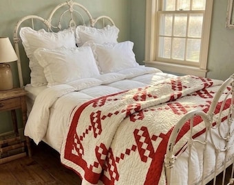 Puede incluir: Dormitorio con una cama de metal blanco y un edredón rojo y blanco estampado. La cama está hecha con ropa de cama y almohadas blancas. Una mesita de noche de madera con una lámpara y libros está al lado de la cama.