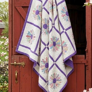 Op de afbeelding: Een witte quilt met een paarse rand en ruitpatroon, versierd met kleurrijke bloemmotieven. De quilt is gedrapeerd over een verweerde rode houten deur, die het ingewikkelde patchwork en de levendige kleuren laat zien.