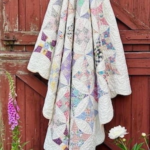 May include: A quilt featuring a white background with a repeating pattern of star-shaped blocks. The blocks are composed of various small floral and geometric fabric prints in shades of pink, blue, yellow, purple, and green, with scalloped edges.