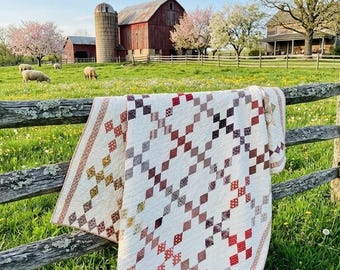 Könnte beinhalten: Ein handgefertigter Quilt mit einem Rautenmuster in Rot-, Braun- und Grautönen ist über einen Holzzaun drapiert. Der Quilt befindet sich vor einer ländlichen Landschaft mit einer roten Scheune, einem Silo und grasenden Schafen.