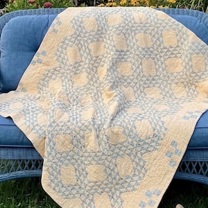 May include: A cream-colored quilt featuring a repeating geometric pattern in light blue. The quilt is constructed with intricate stitching and displays a series of interconnected shapes and small square motifs across its surface.