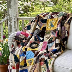 May include: A colorful patchwork quilt draped over a white wicker chair on a porch. The quilt features various fabric squares in shades of red, blue, yellow, and brown, with embroidered details. The background shows a garden with flowers.