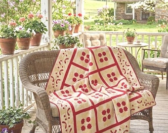 May include: A rectangular quilt featuring a cream-colored background with a repeating grid pattern outlined in red. Each square contains a four-petaled red floral motif, while the border displays curved red shapes and linear accents.