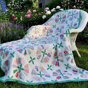 May include: A white quilt featuring a repeating pattern of circular fan motifs made from various colorful patterned fabric scraps, accented with green four-leaf clover shapes and finished with a solid green border draped over a white wicker chair.