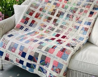 Puede incluir: Un edredón hecho a mano con un diseño de retazos, con cuadrados de varios colores y patrones, incluyendo estampados florales y geométricos. El edredón está sobre un sofá de mimbre blanco, con texto visible. La paleta de colores incluye rojo, azul, rosa y blanco.