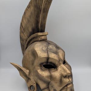 Pode incluir: Uma máscara de cor bronze com uma crista alta e texturizada. A máscara apresenta orelhas pontudas, um nariz proeminente e um design segmentado detalhado. As órbitas oculares são escuras, e a estética geral sugere um tema de fantasia ou histórico.