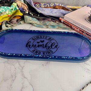 May include: A long, oval-shaped tray with a deep blue interior and a glittery blue border. The tray features the words "ALWAYS BE HUMBLE AND KIND" in black script. The tray is a decorative item, suitable for holding small items.