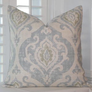 May include: Decorative throw pillow with a light blue and off-white damask pattern. The pillow features a large central medallion design with scrolling details and accents of muted green. The pillow is made of a textured fabric.