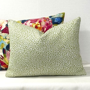 May include: A rectangular decorative pillow with a light green dotted pattern on a white background. Another pillow with a colorful floral design is partially visible behind it. The pillows are on a white surface.