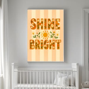 May include: A framed art print with the words "SHINE BRIGHT" in orange letters filled with floral designs. The artwork features a yellow sun and floral accents on a striped background, suitable for a nursery or children's room.