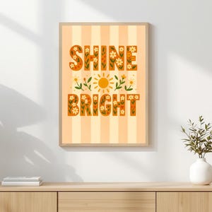 May include: A framed art print with the words "SHINE BRIGHT" in orange floral lettering. The background is striped in light orange and white. A yellow sun and floral accents are in the center. The artwork is displayed on a wooden cabinet.