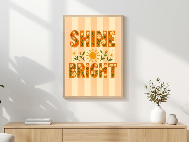 May include: A framed art print with the words "SHINE BRIGHT" in orange floral lettering. The background is striped in light orange and white. A yellow sun and floral accents are in the center. The artwork is displayed on a wooden cabinet.