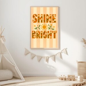 May include: A framed art print with the words "Shine Bright" in orange floral letters. The background is striped in light orange and white. The print is displayed on a white wall, with a teepee and other decor in the foreground.