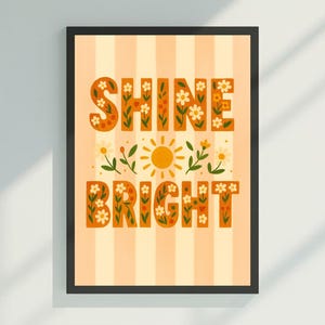 May include: Framed art print with the words "SHINE BRIGHT" in orange, floral-decorated letters. The background is striped in light orange and white. A yellow sun and floral accents are between the words. The frame is black.