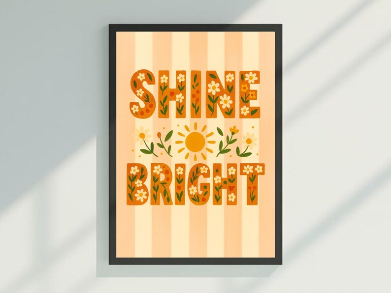 May include: Framed art print with the words "SHINE BRIGHT" in orange, floral-decorated letters. The background is striped in light orange and white. A yellow sun and floral accents are between the words. The frame is black.