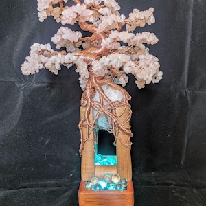 May include: A handcrafted bonsai tree sculpture with a copper wire trunk and branches, adorned with pink gemstone leaves. The tree sits atop a wooden base with a glowing blue light feature and clear glass marbles.