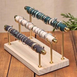 Puede incluir: Un soporte de anillos de madera con tres varillas de metal dorado. Cada varilla está cubierta con un tejido de un color diferente y tiene anillos apilados en ella. El soporte está sobre una superficie de madera.