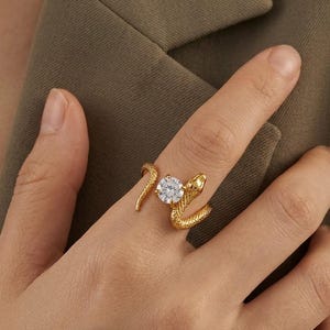 Puede incluir: Un anillo de tono dorado con forma de serpiente enroscada con una superficie texturizada similar a escamas. El cuerpo de la serpiente envuelve una única piedra preciosa redonda, transparente y facetada, creando un diseño abierto.