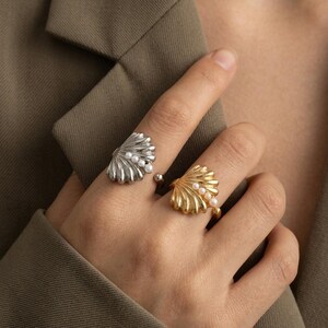 Könnte beinhalten: Zwei offene Ringe in Form von fächerartigen Muscheln. Ein Ring ist silberfarben, der andere goldfarben. Jeder Ring ist mit einer Reihe kleiner, runder weißer Perlen besetzt, die mittig in das muschelförmige Design eingearbeitet sind.