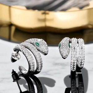 May include: A pair of silver-toned snake earrings. Each earring features a coiled snake design encrusted with small, clear crystals. The snake's eye is a small, green gemstone. The earrings have a post and backing for secure wear.