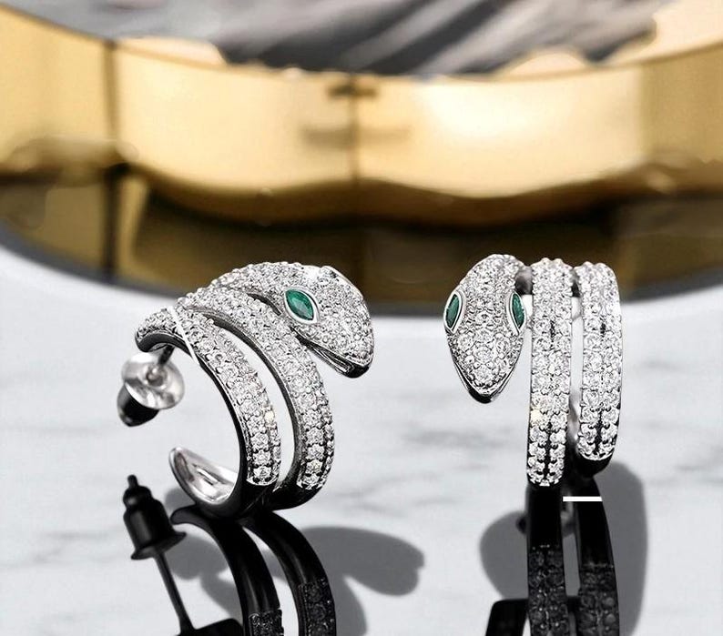 May include: A pair of silver-toned snake earrings. Each earring features a coiled snake design encrusted with small, clear crystals. The snake's eye is a small, green gemstone. The earrings have a post and backing for secure wear.