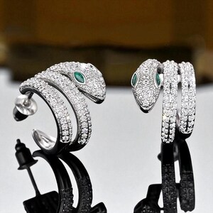 May include: A pair of silver-toned snake earrings. Each earring features a snake design encrusted with small, clear crystals. The snake's eyes are small, oval-shaped green stones. The earrings are displayed against a reflective surface.