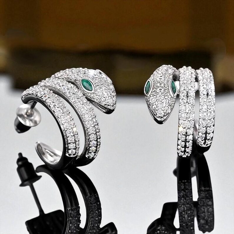 May include: A pair of silver-toned snake earrings. Each earring features a snake design encrusted with small, clear crystals. The snake's eyes are small, oval-shaped green stones. The earrings are displayed against a reflective surface.
