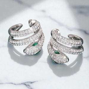 May include: Two silver-toned snake-shaped ear cuffs. Each cuff is encrusted with small, clear crystals and features a small, green gemstone for the eye. The snake design wraps around the ear, creating a unique and elegant look. The background is a light marble.