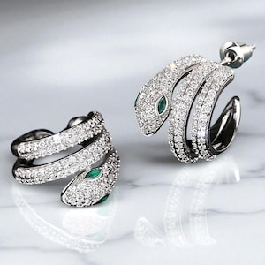 May include: A pair of silver-toned snake earrings encrusted with clear crystals. The snake's head features a small, green gemstone eye. The earrings are designed to wrap around the ear, creating a unique and elegant look.