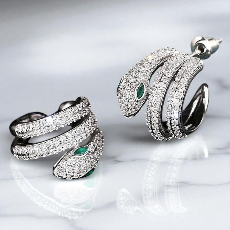 May include: A pair of silver-toned snake earrings encrusted with clear crystals. The snake's head features a small, green gemstone eye. The earrings are designed to wrap around the ear, creating a unique and elegant look.