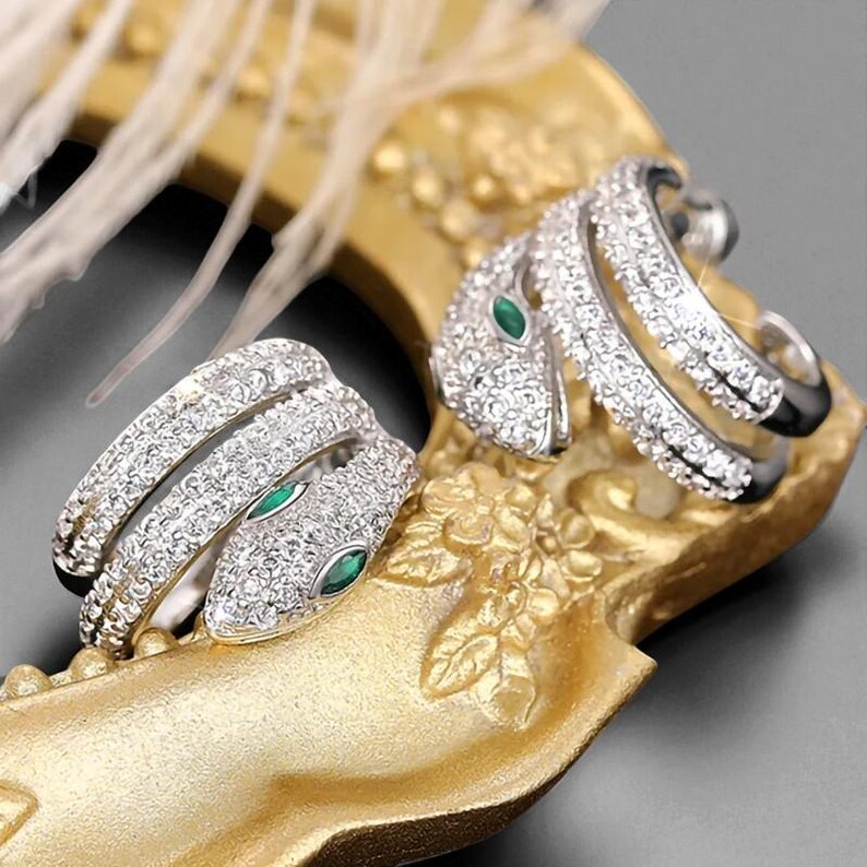 May include: Two silver snake rings encrusted with clear crystals and emerald green eyes. The rings are displayed on a gold-colored decorative tray with floral detailing. The rings are a unique jewelry item.