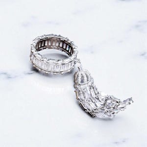 May include: A silver ring with a band of baguette-cut stones and a dangling tassel pendant. The tassel has a crown-like top and rectangular stones. The ring and pendant are set against a white background.