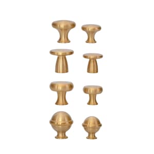 May include: A collection of eight brass cabinet knobs in various shapes. The knobs include mushroom, cylindrical, and spherical designs. The hardware is displayed against a white background, showcasing the warm, metallic finish of the knobs.