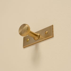 May include: A brass wall hook with a round knob and rectangular backplate. The hook is mounted on a cream-coloured wall. The hardware is made of brass and has a matte finish. The hook is designed for hanging items.