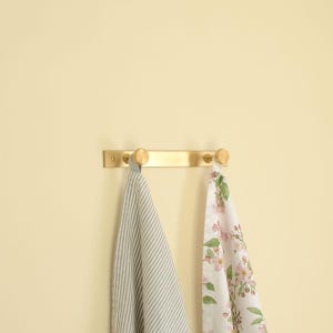 May include: A brass towel rail with three round knobs, mounted on a light yellow wall. Two decorative towels hang from the rail: one with grey and white stripes, and the other with a floral pattern.
