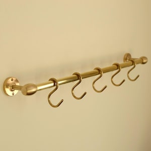 May include: A brass-coloured wall-mounted rack with six curved hooks. The rack has a cylindrical bar and is attached to the wall with round, brass-coloured mounting hardware. The hooks are evenly spaced along the bar.