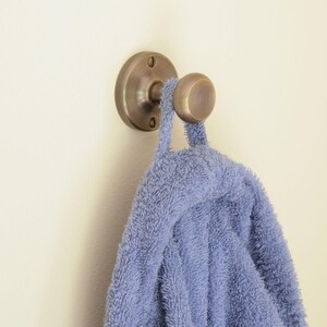 May include: A bronze-coloured wall hook with a round knob and a circular base, mounted on a cream-coloured wall. A blue towel is hanging from the hook. The hook is designed for hanging towels or robes in a bathroom.